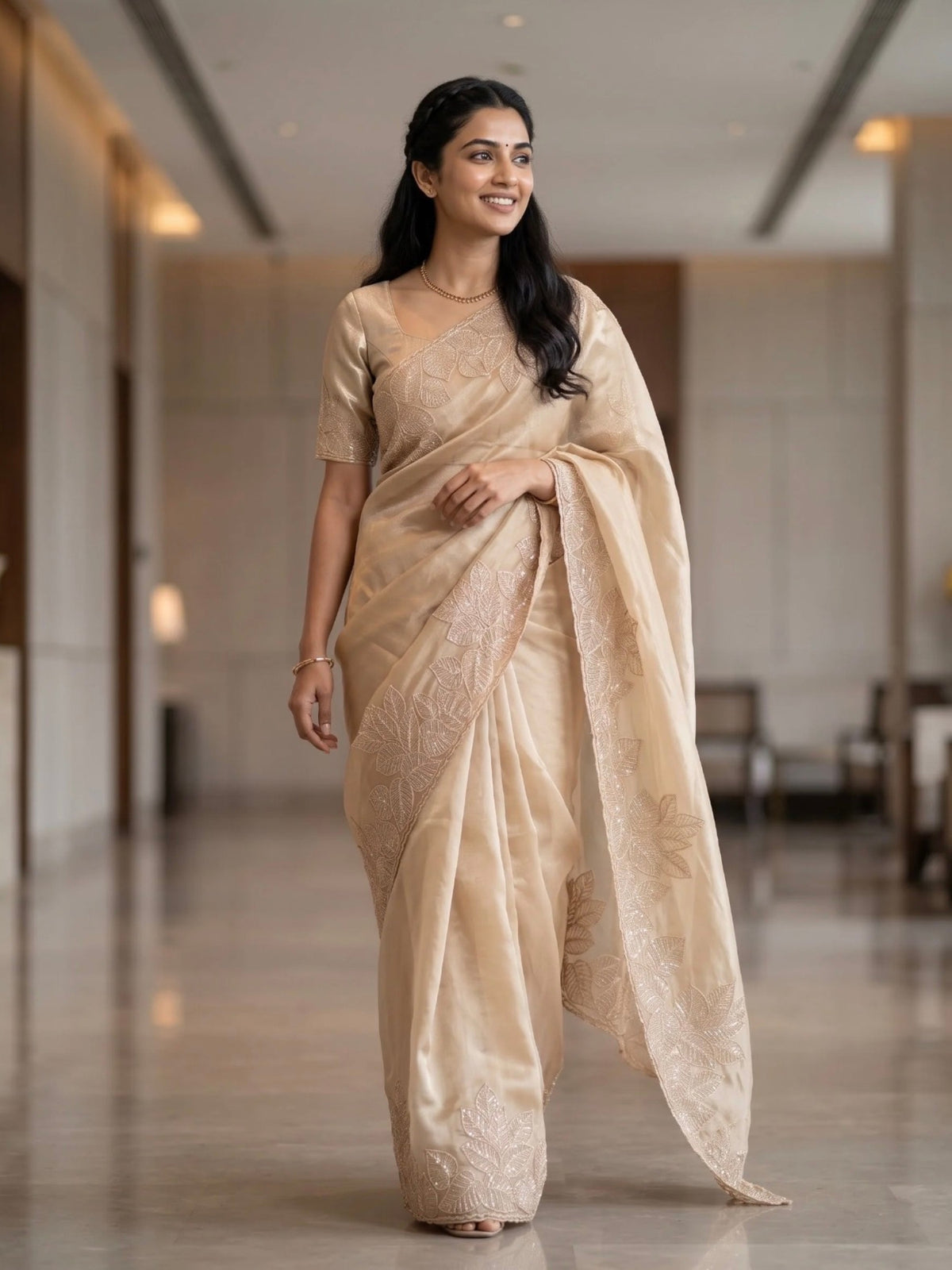Woman wearing a beige saree with subtle floral embroidery and a matching blouse indoors.