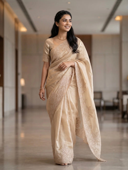 Woman wearing a beige saree with subtle floral embroidery and a matching blouse indoors.