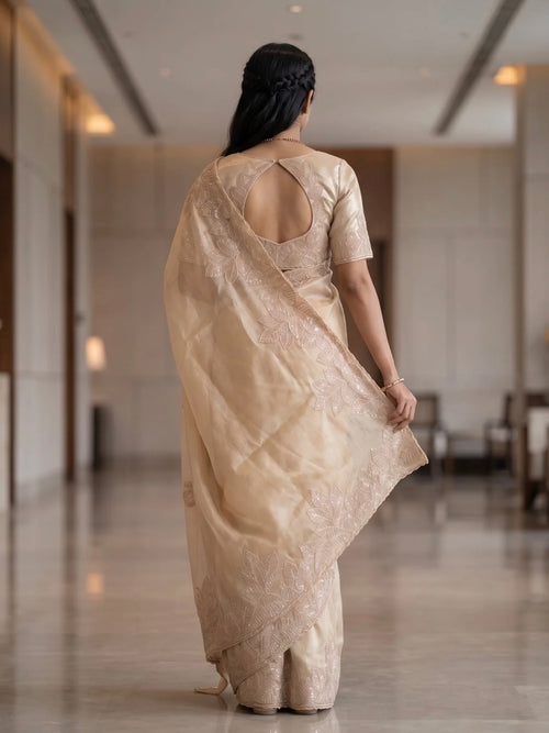 Woman wearing a beige saree with subtle floral embroidery and a matching blouse indoors.