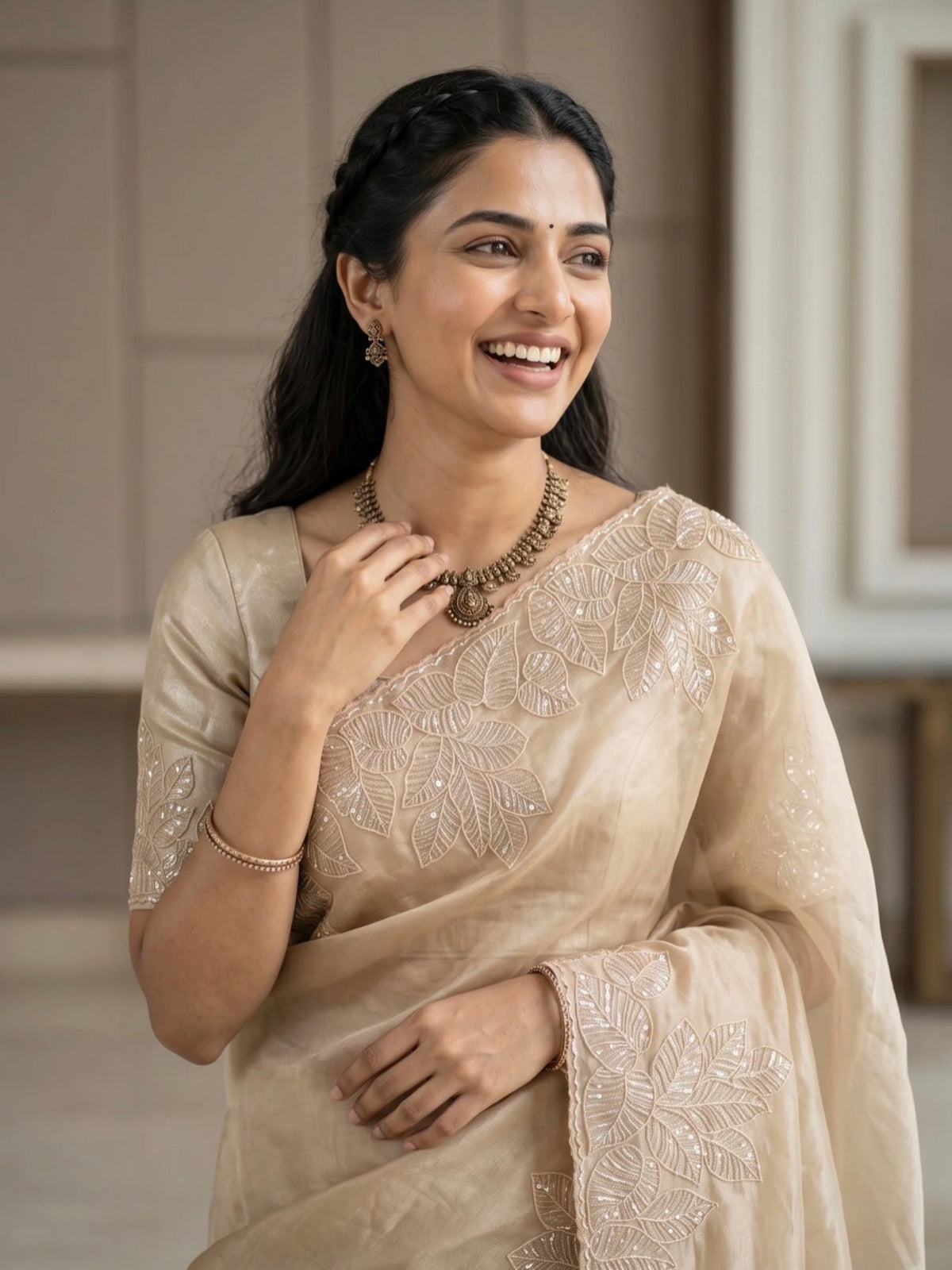 Woman wearing a beige saree with subtle floral embroidery and a matching blouse indoors.