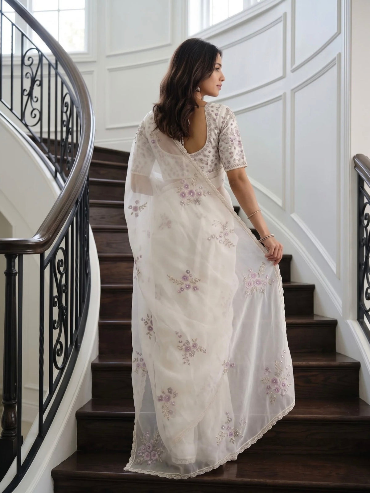 Woman wearing a white saree with lavender floral prints, standing on an elegant staircase.