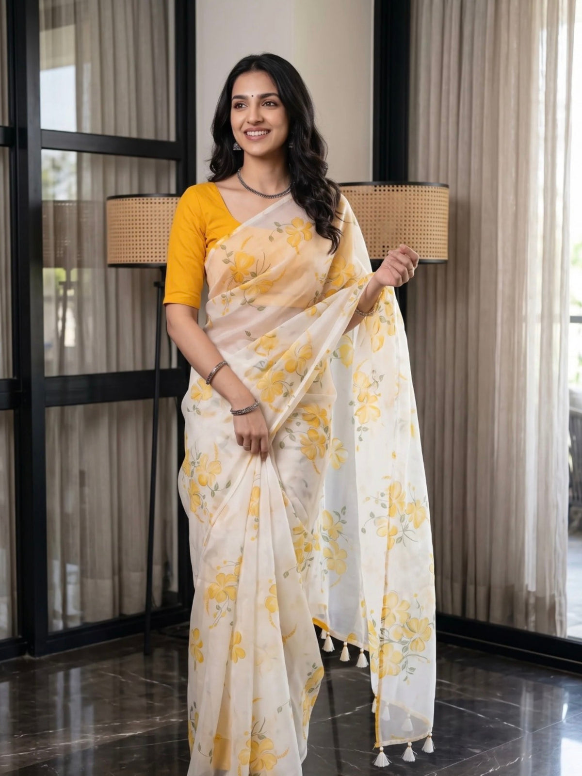 Woman wearing a white saree with yellow floral prints and tassel details indoors.