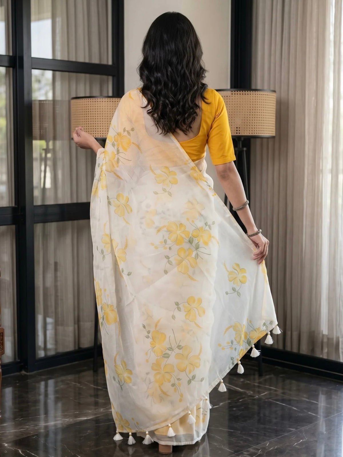Woman wearing a white saree with yellow floral prints and tassel details indoors.