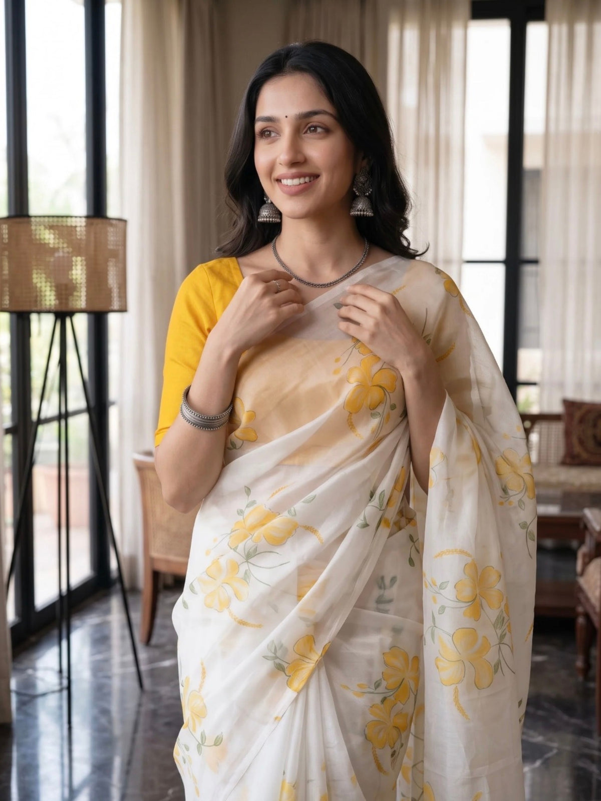 Woman wearing a white saree with yellow floral prints and tassel details indoors.