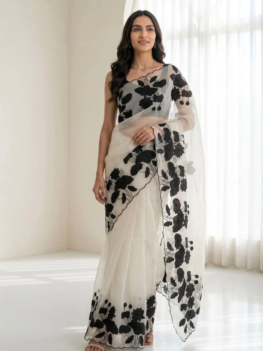 Woman wearing a white saree with black floral prints and a sheer pallu indoors.