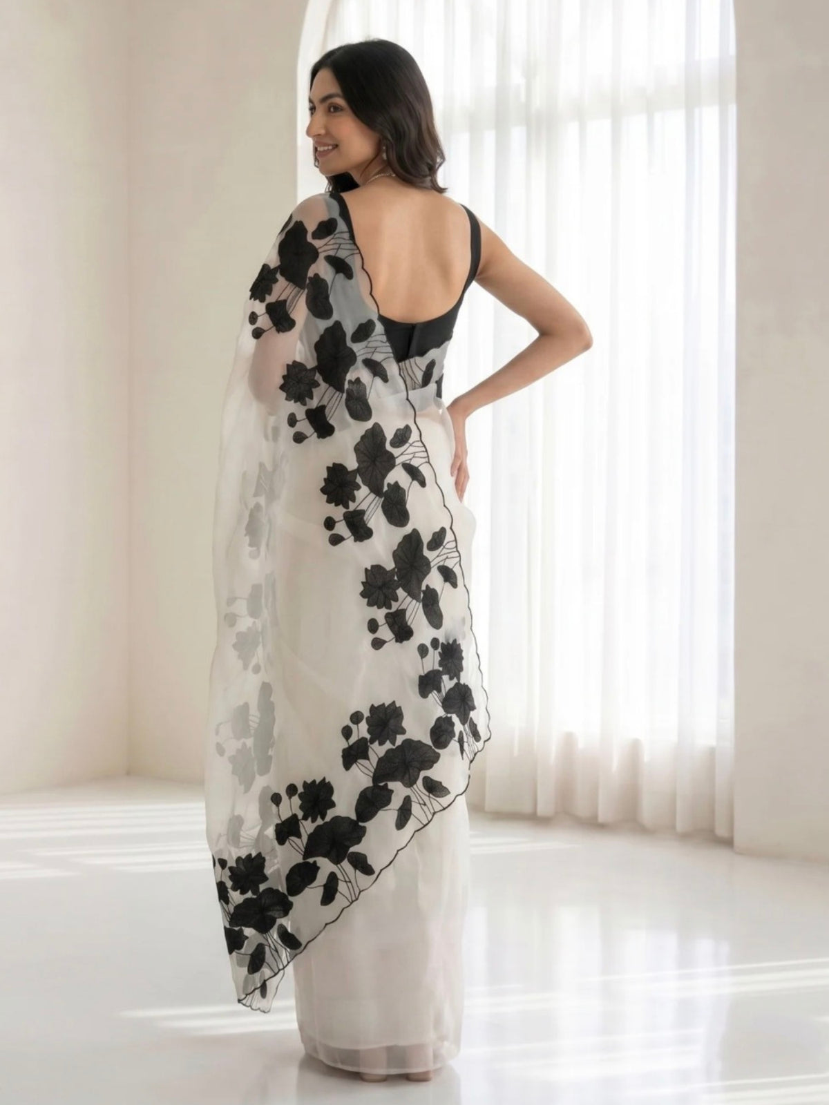 Woman wearing a white saree with black floral prints and a sheer pallu indoors.