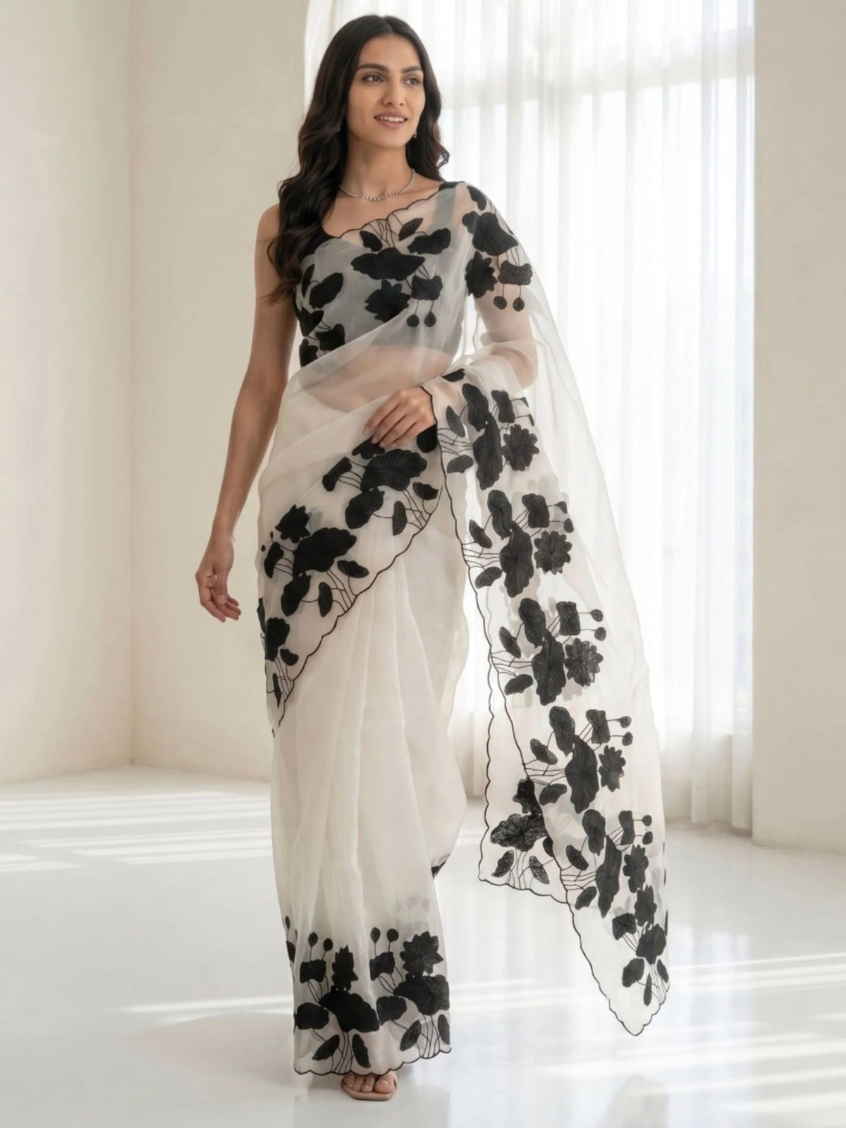 Woman wearing a white saree with black floral prints and a sheer pallu indoors.