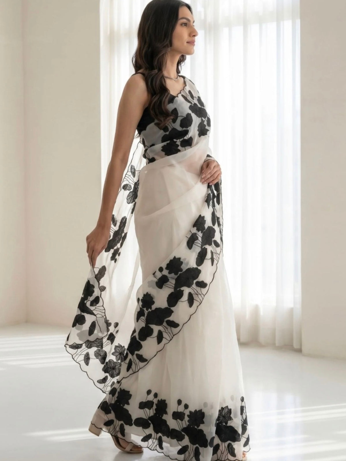 Woman wearing a white saree with black floral prints and a sheer pallu indoors.