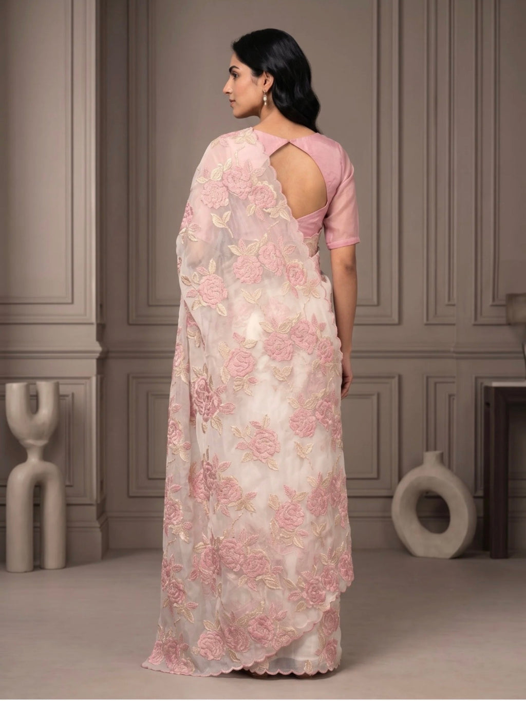 Woman wearing a white saree with pink floral embroidery and a matching pink blouse indoors.