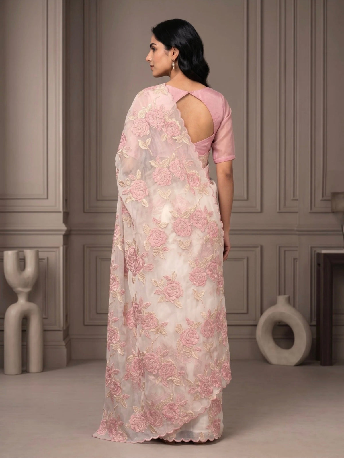 Woman wearing a white saree with pink floral embroidery and a matching pink blouse indoors.