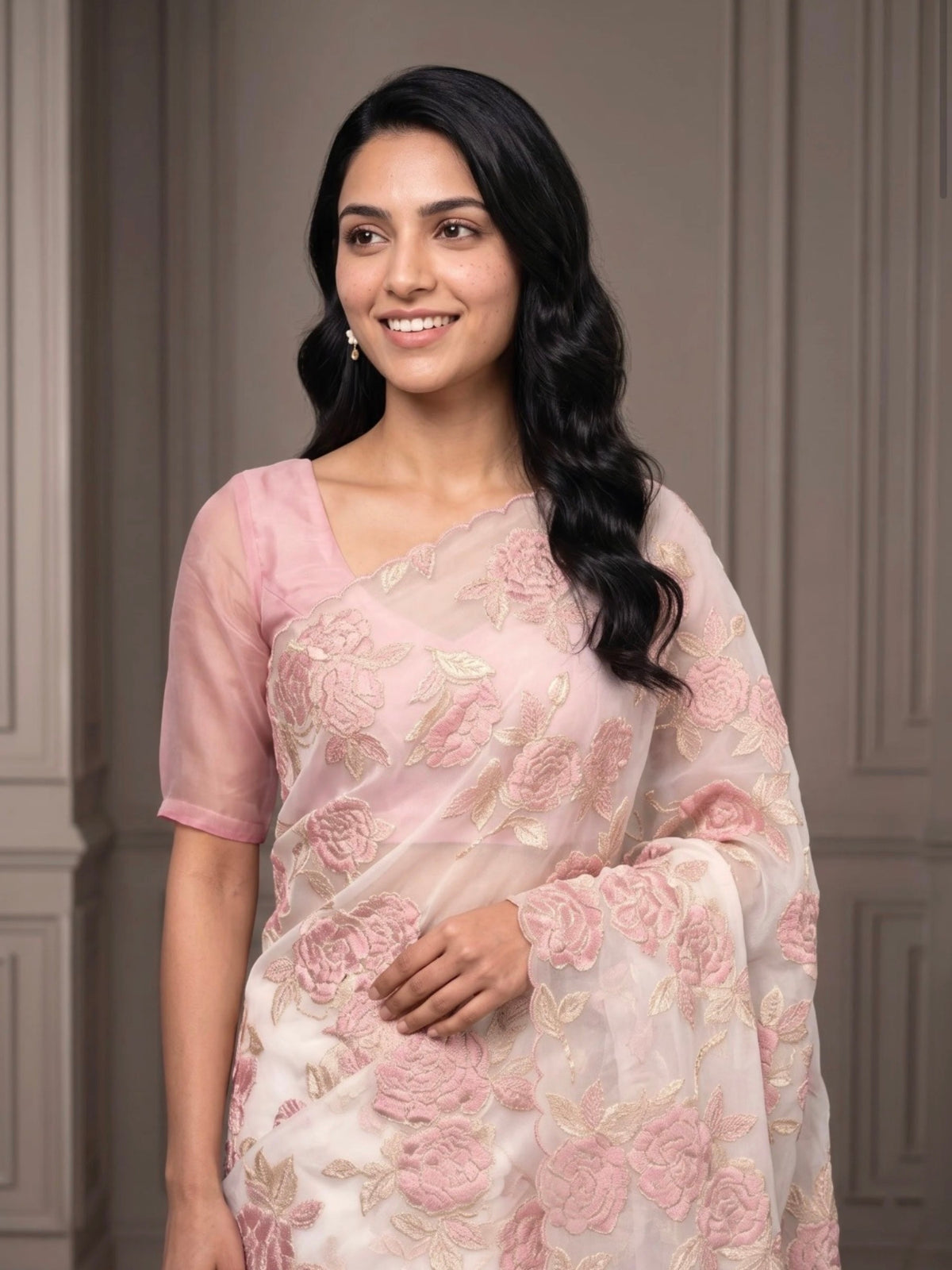 Woman wearing a white saree with pink floral embroidery and a matching pink blouse indoors.
