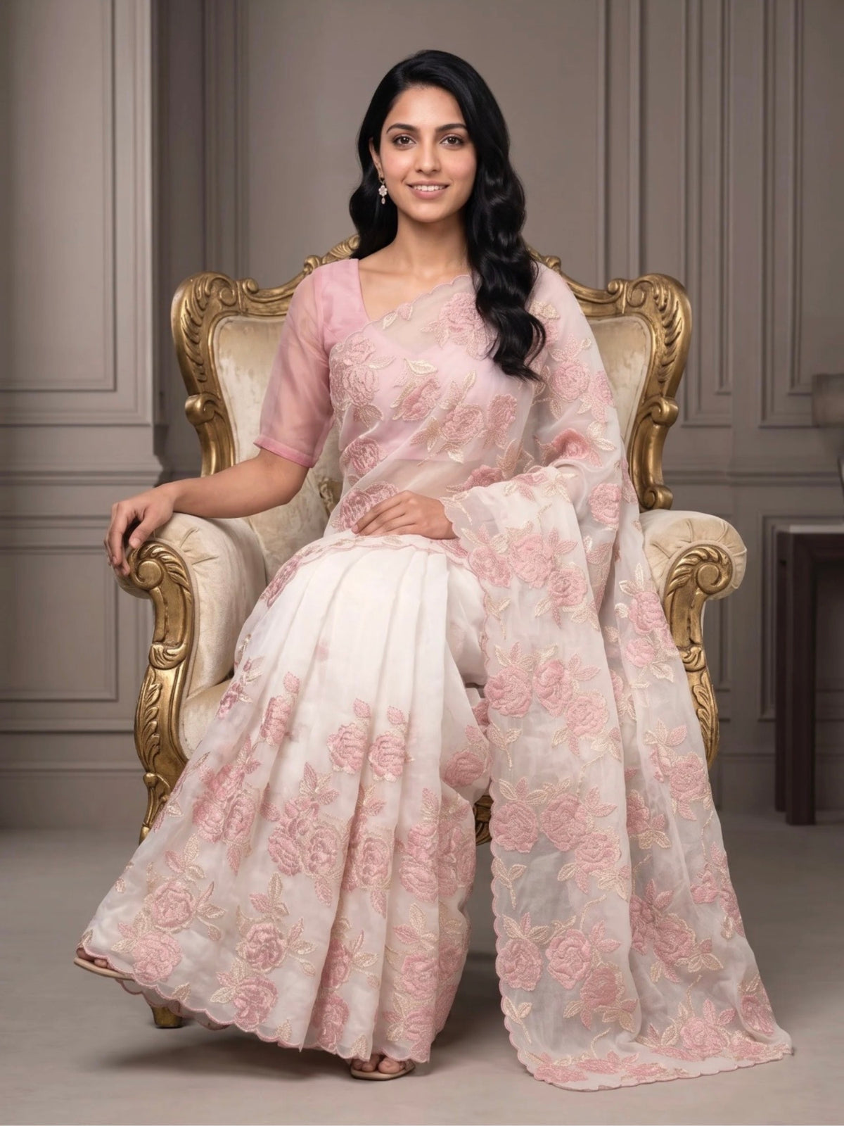 Woman wearing a white saree with pink floral embroidery and a matching pink blouse indoors.